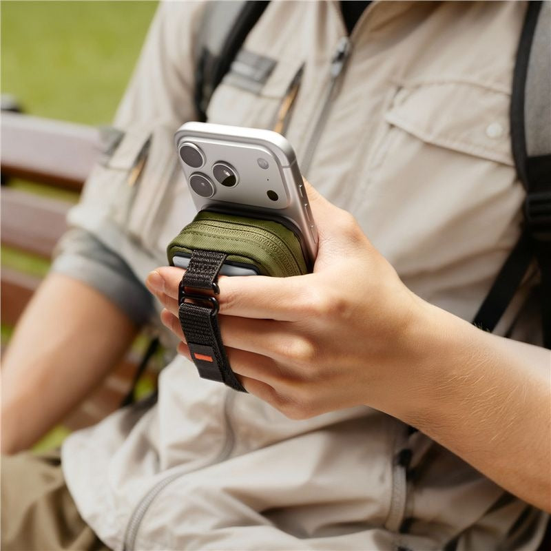 A close-up of a person holding a silver smartphone with an olive green and black phone strap accessory attached to the camera area, seated outdoors on a bench.