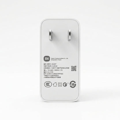 A white Xiaomi power adapter with a white plug, shown from the back on a clean white background. The back features black printed specifications, a QR code, and various certification logos.